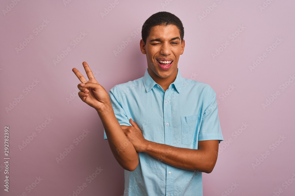 Young handsome arab man wearing blue shirt standing over isolated pink background smiling with happy face winking at the camera doing victory sign. Number two. Young handsome arab man wearing blue shirt standing over isolated pink background smiling with happy face winking at the camera doing victory sign. Number two.
