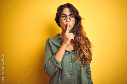 Young beautiful woman wearing green shirt and glasses over yelllow isolated background asking to be quiet with finger on lips. Silence and secret concept.