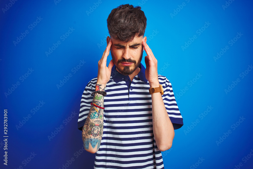 Young man with tattoo wearing striped polo standing over isolated blue background with hand on head for pain in head because stress. Suffering migraine. Young man with tattoo wearing striped polo standing over isolated blue background with hand on head for pain in head because stress. Suffering migraine.