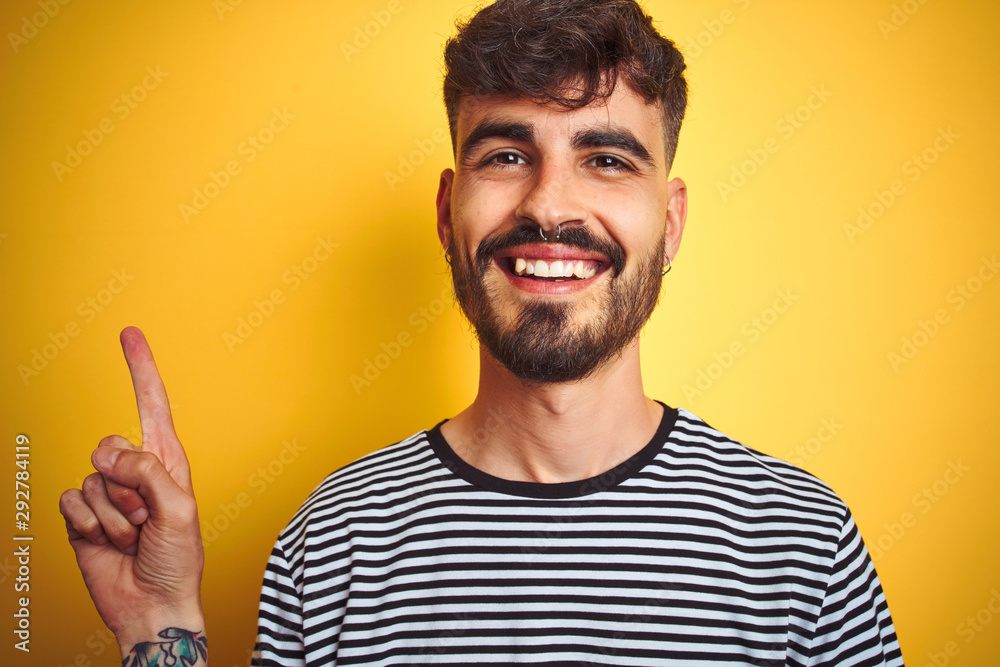 Young man with tattoo wearing striped t-shirt standing over isolated yellow background with a big smile on face, pointing with hand and finger to the side looking at the camera. Young man with tattoo wearing striped t-shirt standing over isolated yellow background with a big smile on face, pointing with hand and finger to the side looking at the camera.