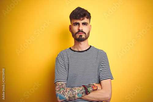 Young man with tattoo wearing striped t-shirt standing over isolated yellow background skeptic and nervous, disapproving expression on face with crossed arms. Negative person.