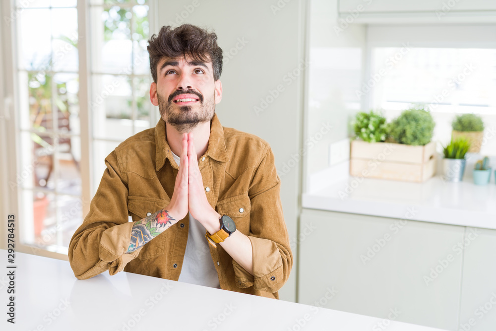 Young man wearing casual jacket sitting on white table begging and praying with hands together with hope expression on face very emotional and worried. Asking for forgiveness. Religion concept.
