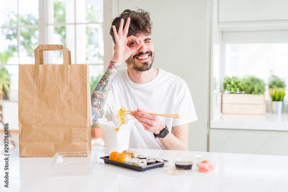 Young man eating asian sushi from home delivery with happy face smiling doing ok sign with hand on eye looking through fingers Young man eating asian sushi from home delivery with happy face smiling doing ok sign with hand on eye looking through fingers