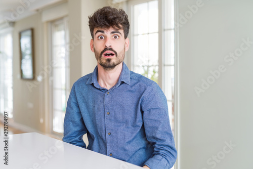 Young man wearing casual shirt sitting on white table afraid and shocked with surprise expression, fear and excited face.