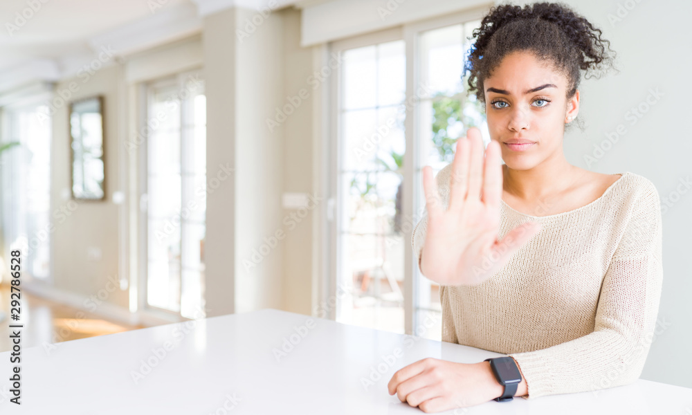 Beautiful young african american woman with afro hair sitting on table at home doing stop sing with palm of the hand. Warning expression with negative and serious gesture on the face. Beautiful young african american woman with afro hair sitting on table at home doing stop sing with palm of the hand. Warning expression with negative and serious gesture on the face.