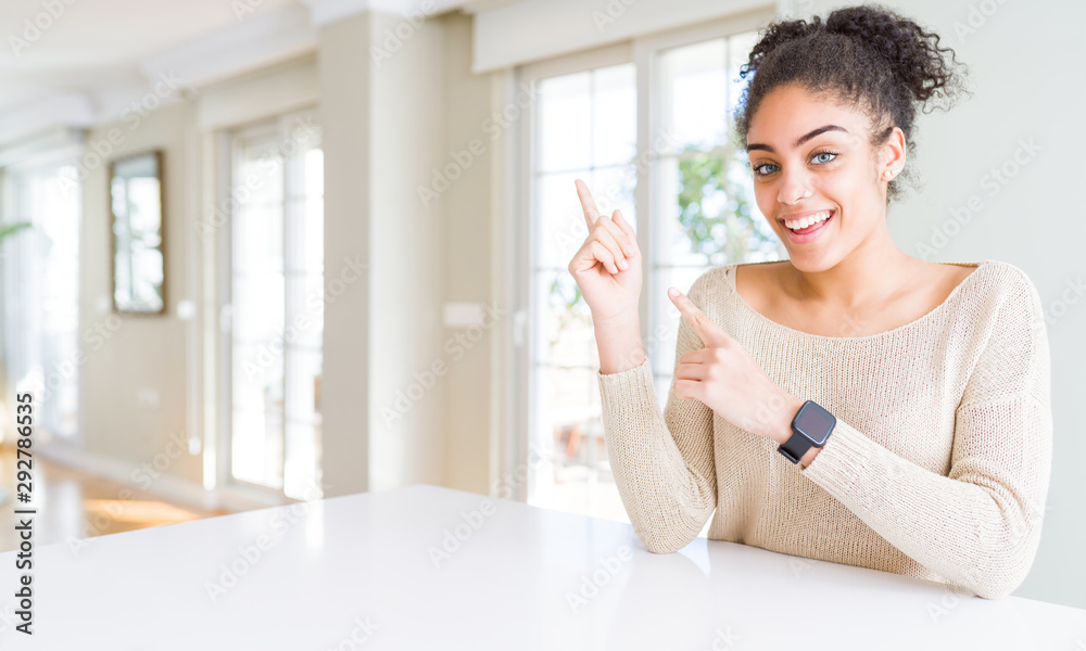 Beautiful young african american woman with afro hair sitting on table at home smiling and looking at the camera pointing with two hands and fingers to the side. Beautiful young african american woman with afro hair sitting on table at home smiling and looking at the camera pointing with two hands and fingers to the side.