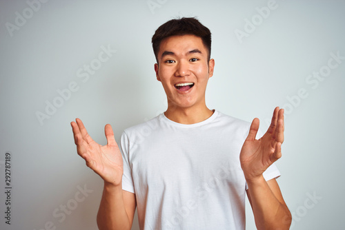 Young asian chinese man wearing t-shirt standing over isolated white background celebrating crazy and amazed for success with arms raised and open eyes screaming excited. Winner concept