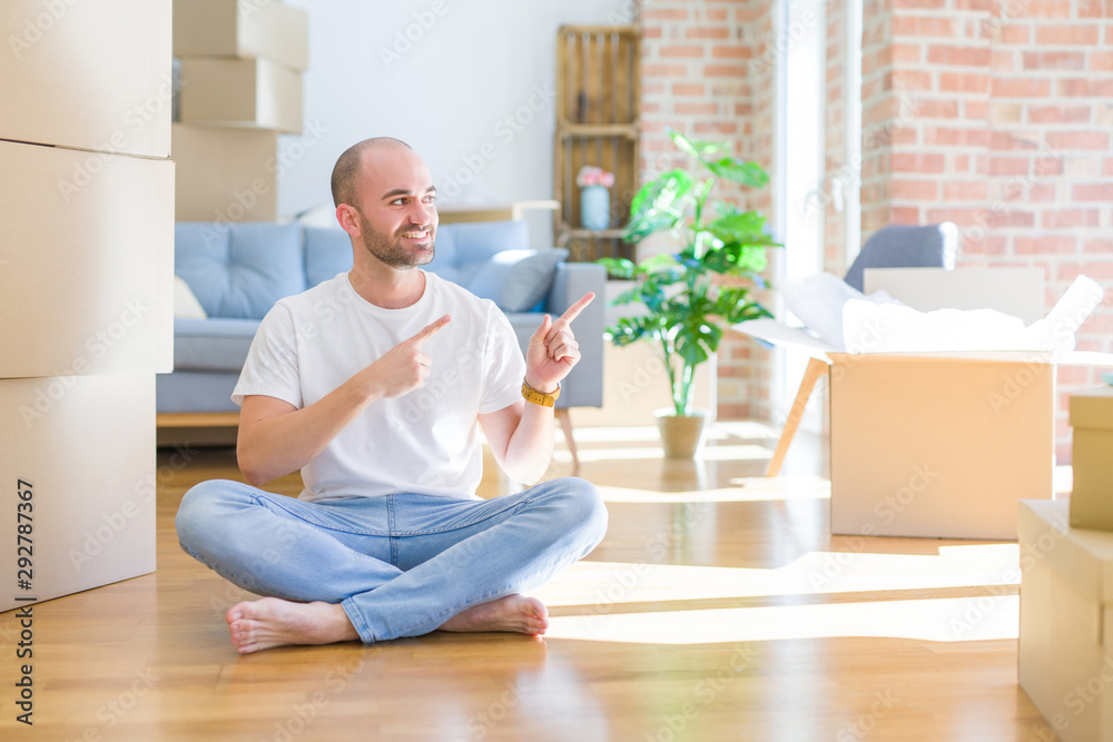 Young bald man sitting on the floor around cardboard boxes moving to a new home smiling and looking at the camera pointing with two hands and fingers to the side.