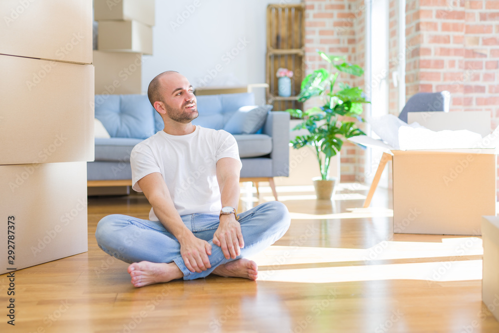 Young bald man sitting on the floor around cardboard boxes moving to a new home looking away to side with smile on face, natural expression. Laughing confident. Young bald man sitting on the floor around cardboard boxes moving to a new home looking away to side with smile on face, natural expression. Laughing confident.