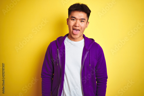 Young asian chinese man wearing purple sweatshirt standing over isolated yellow background sticking tongue out happy with funny expression. Emotion concept.