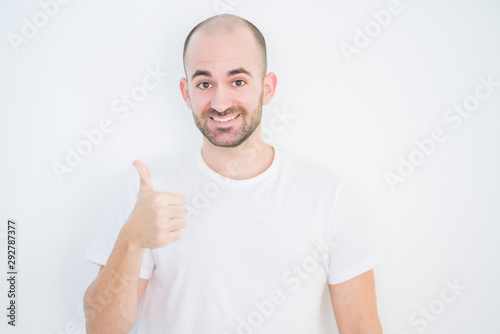 Young bald man over white isolated background doing happy thumbs up gesture with hand. Approving expression looking at the camera with showing success.