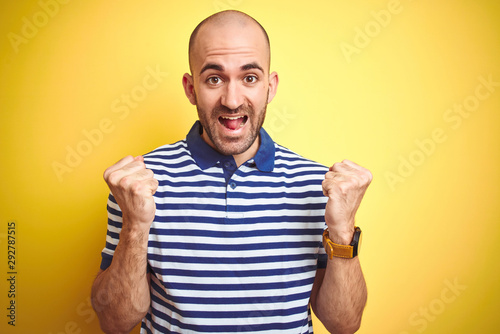 Young bald man with beard wearing casual striped blue t-shirt over yellow isolated background celebrating surprised and amazed for success with arms raised and open eyes. Winner concept.