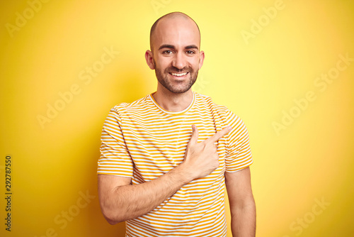Young bald man with beard wearing casual striped t-shirt over yellow isolated background cheerful with a smile on face pointing with hand and finger up to the side with happy and natural expression