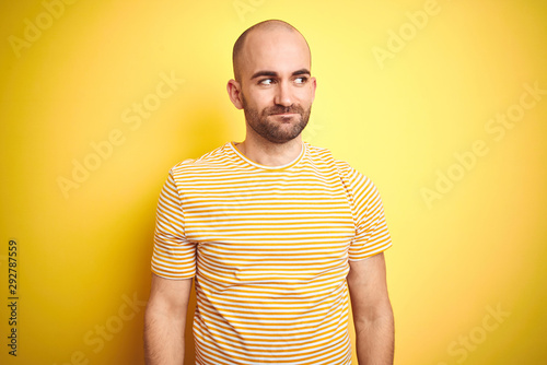 Young bald man with beard wearing casual striped t-shirt over yellow isolated background smiling looking to the side and staring away thinking.