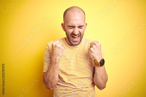Young bald man with beard wearing casual striped t-shirt over yellow isolated background excited for success with arms raised and eyes closed celebrating victory smiling. Winner concept.