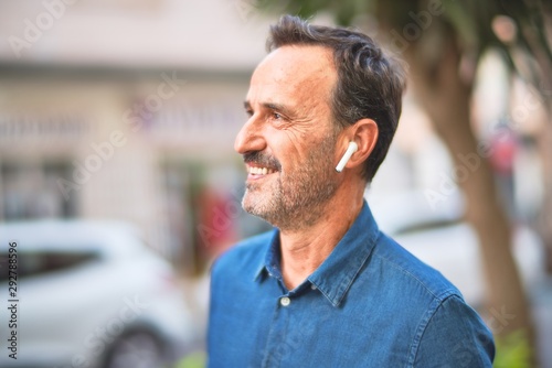 Middle age handsome businessman standing on the street listening to music using earphones smiling