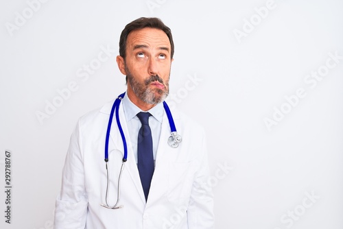 Middle age doctor man wearing coat and stethoscope standing over isolated white background making fish face with lips, crazy and comical gesture. Funny expression. © Krakenimages.com