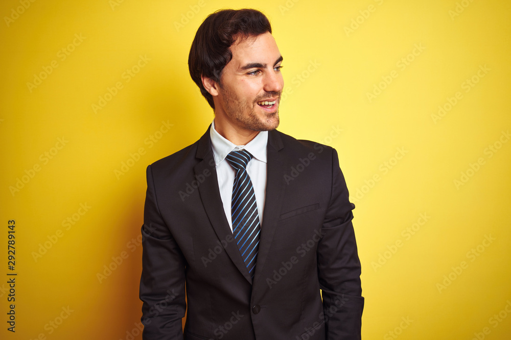 Young handsome businessman wearing suit and tie standing over isolated yellow background looking away to side with smile on face, natural expression. Laughing confident. Young handsome businessman wearing suit and tie standing over isolated yellow background looking away to side with smile on face, natural expression. Laughing confident.
