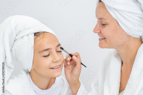 Happy family at home. Mother and young daughter while applying makeup at home. Mom and child girl are in bathrobes and with towels on their heads