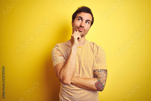 Young handsome man with tattoo wearing striped t-shirt over isolated yellow background with hand on chin thinking about question, pensive expression. Smiling with thoughtful face. Doubt concept. © Krakenimages.com