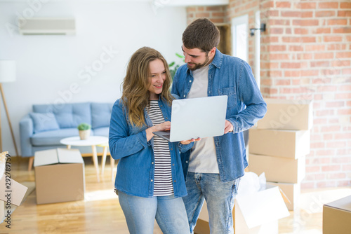 Young couple using computer laptop standing on a room around cardboard boxes, happy for moving to a new apartment