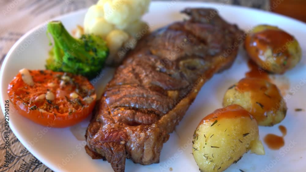 slow stabilized shot of grilled steak on white plate with grilled tomato, broccoli, cauliflower and baked potatoes slow stabilized shot of grilled steak on white plate with grilled tomato, broccoli, cauliflower and baked potatoes