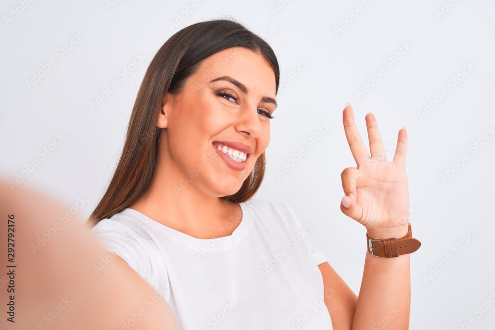 Beautiful young woman taking a selfie photo using smartphone over white background doing ok sign with fingers, excellent symbol Beautiful young woman taking a selfie photo using smartphone over white background doing ok sign with fingers, excellent symbol