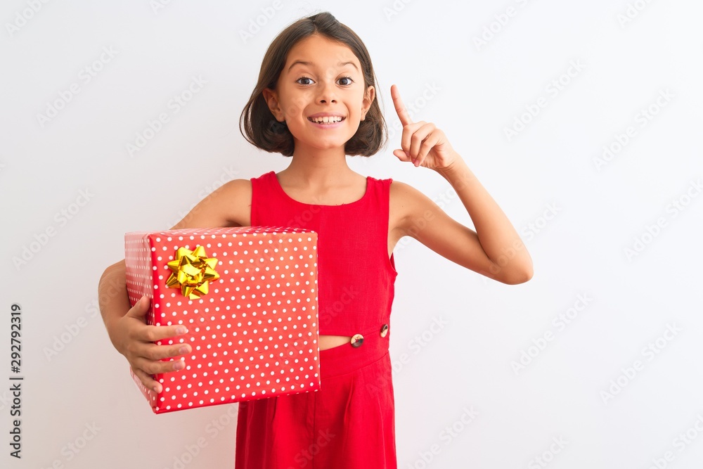 Beautiful child girl holding birthday gift standing over isolated white background surprised with an idea or question pointing finger with happy face, number one Beautiful child girl holding birthday gift standing over isolated white background surprised with an idea or question pointing finger with happy face, number one