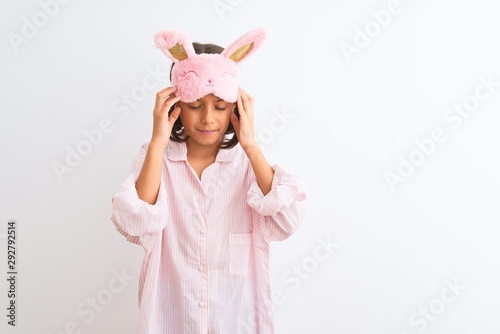 Beautiful child girl wearing sleep mask and pajama standing over isolated white background with hand on head for pain in head because stress. Suffering migraine.