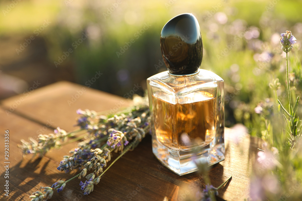 Bottle of luxury perfume and lavender flowers on wooden table in blooming field. Space for text - obrazy, fototapety, plakaty 