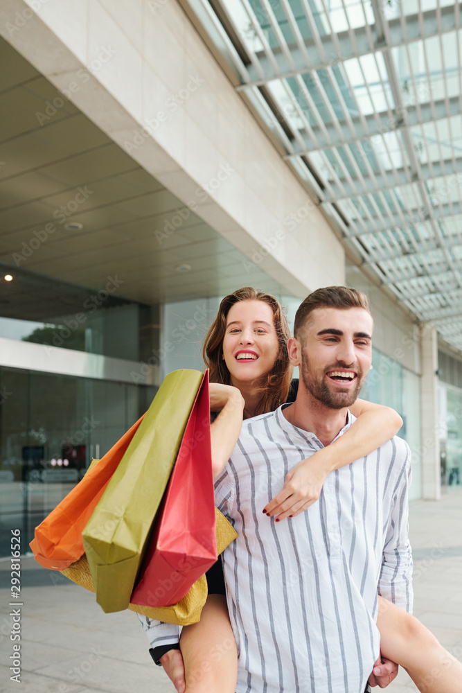 Attractive cheerful young man giving piggyback ride to his lovely girlfriend with many colorful shopping bags when standing at mall Attractive cheerful young man giving piggyback ride to his lovely girlfriend with many colorful shopping bags when standing at mall