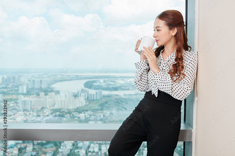 Pensive young pretty entrepreneur standing at big office window, enjoying cup coffee and city view
