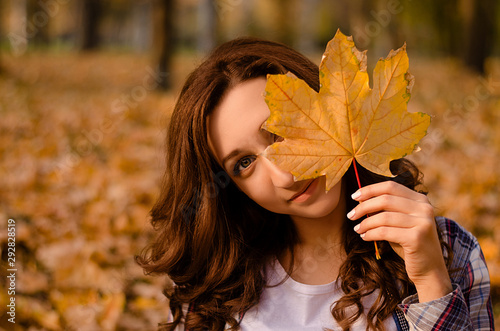 Happy young attractive woman with curly hair is standing in the park and covering her face with yellow autumn maple leaves. Cute girl enjoying warm weather. Autumn mood. Enjoy season. Copy space
