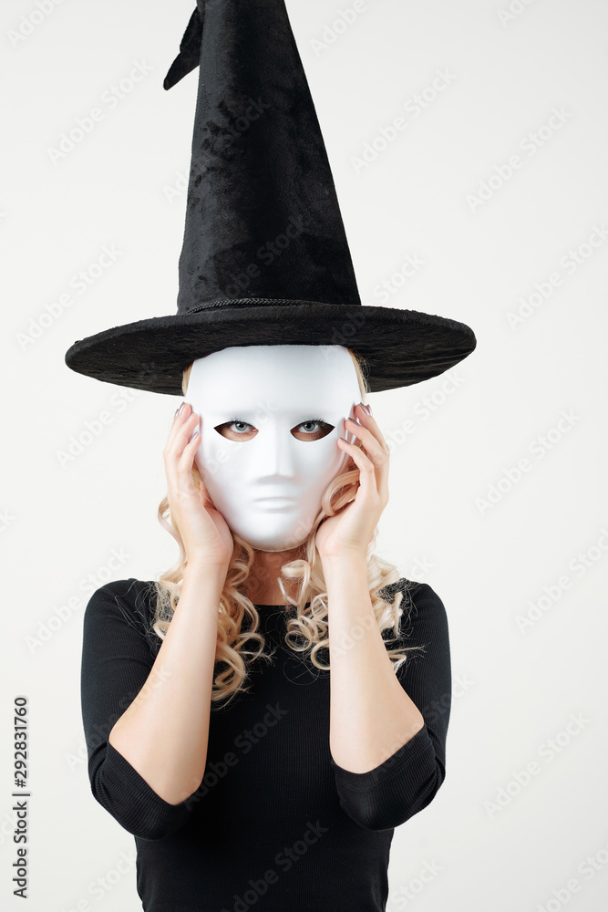 Portrait of young woman in witch costume putting on plastic white mask for Halloween party and looking at camera Portrait of young woman in witch costume putting on plastic white mask for Halloween party and looking at camera