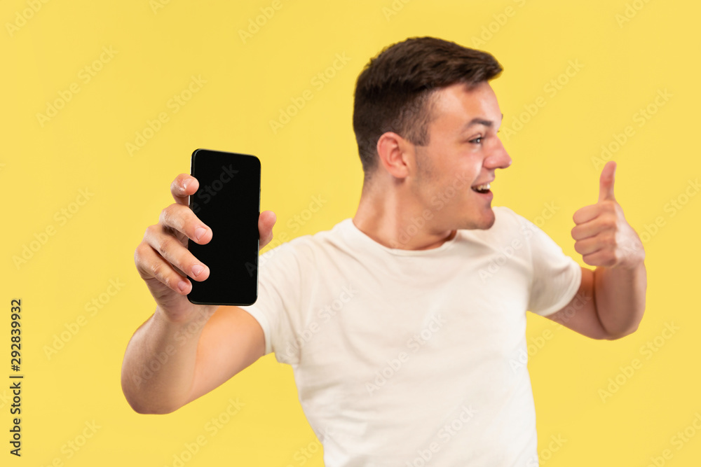 Caucasian young man's half-length portrait on yellow studio background. Beautiful male model in shirt. Concept of human emotions, facial expression, sales, ad. Showing phone's screen and smiling. Caucasian young man's half-length portrait on yellow studio background. Beautiful male model in shirt. Concept of human emotions, facial expression, sales, ad. Showing phone's screen and smiling.