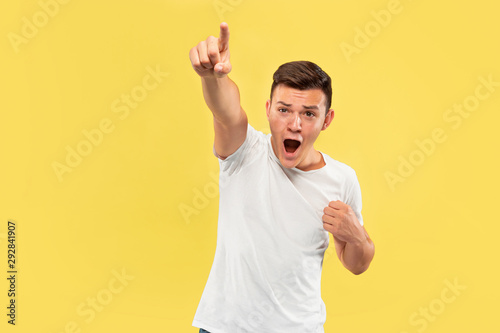 Caucasian young man's half-length portrait on yellow studio background. Beautiful male model in shirt. Concept of human emotions, facial expression, sales, ad. Pointing on and looks happy.