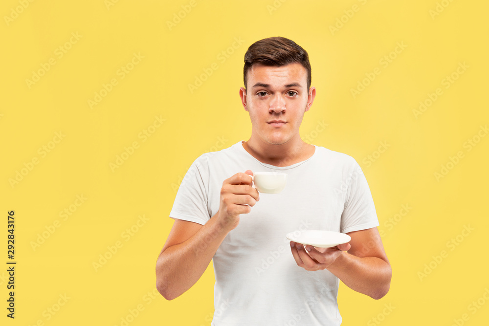 Caucasian young man's half-length portrait on yellow studio background. Beautiful male model in shirt. Concept of human emotions, facial expression, sales, ad. Enjoying drinking coffee or tea. Caucasian young man's half-length portrait on yellow studio background. Beautiful male model in shirt. Concept of human emotions, facial expression, sales, ad. Enjoying drinking coffee or tea.
