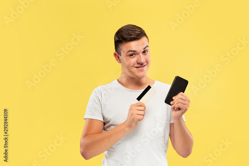 Caucasian young man's half-length portrait on yellow studio background. Beautiful male model in shirt. Concept of human emotions, facial expression, sales, ad. Phone and payment card, online purchases