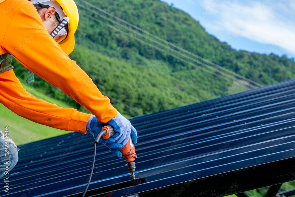 Roofer worker in protective uniform wear and gloves, using air or pneumatic nail gun and installing asphalt shingle on top of the new roof,Concept of residential building under construction.