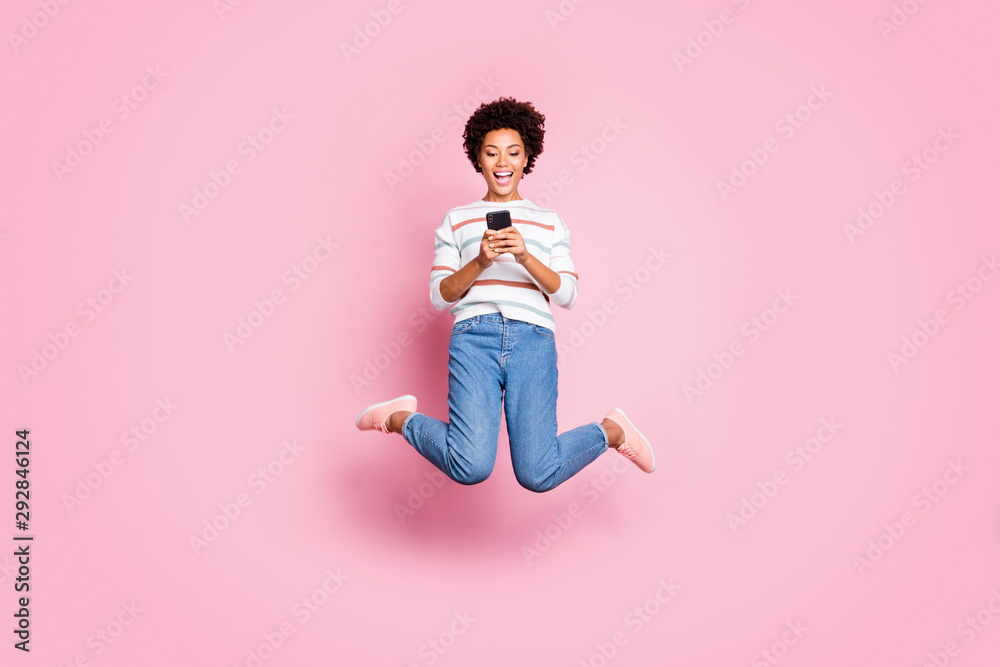 Full length body size photo of cheerful fascinating rejoicing cute girl browsing through her telephone jumping wearing jeans denim striped sweater isolated over pastel color background