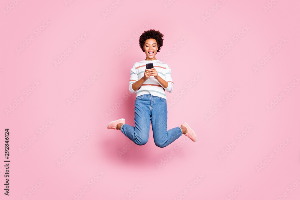 Full length body size photo of cheerful fascinating rejoicing cute girl browsing through her telephone jumping wearing jeans denim striped sweater isolated over pastel color background Full length body size photo of cheerful fascinating rejoicing cute girl browsing through her telephone jumping wearing jeans denim striped sweater isolated over pastel color background