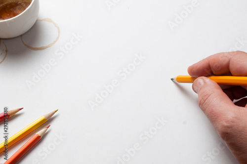 A man hold a pencil over a white sheet of paper, pencils, offee cup and coffee stains as decoration, top view