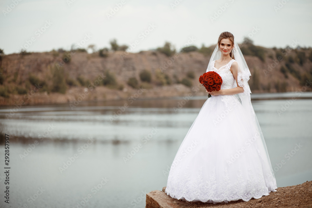 A young smiling and beautiful bride in a white dress is standing against a background of rocks, cliffs and stones. Wedding portrait of a cute curly blonde with a bouquet. Top vew. Space for text.