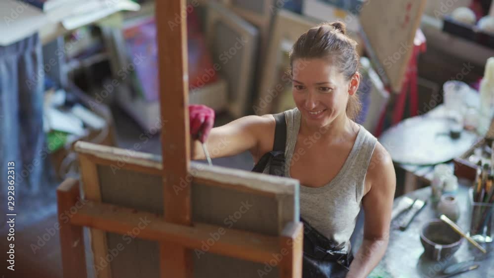 Tracking shot of smiling young female artist enjoying painting on canvas standing at easel in art studio Tracking shot of smiling young female artist enjoying painting on canvas standing at easel in art studio