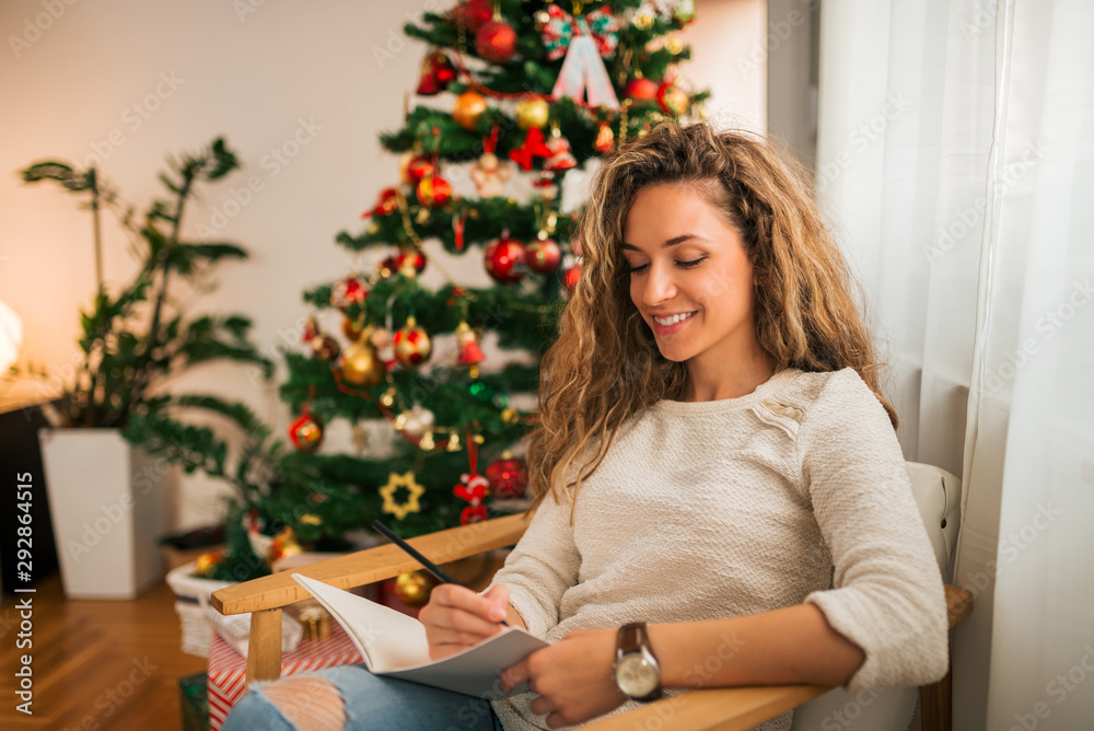 Young positive woman writing in note book near Christmas tree indoors, portrait.