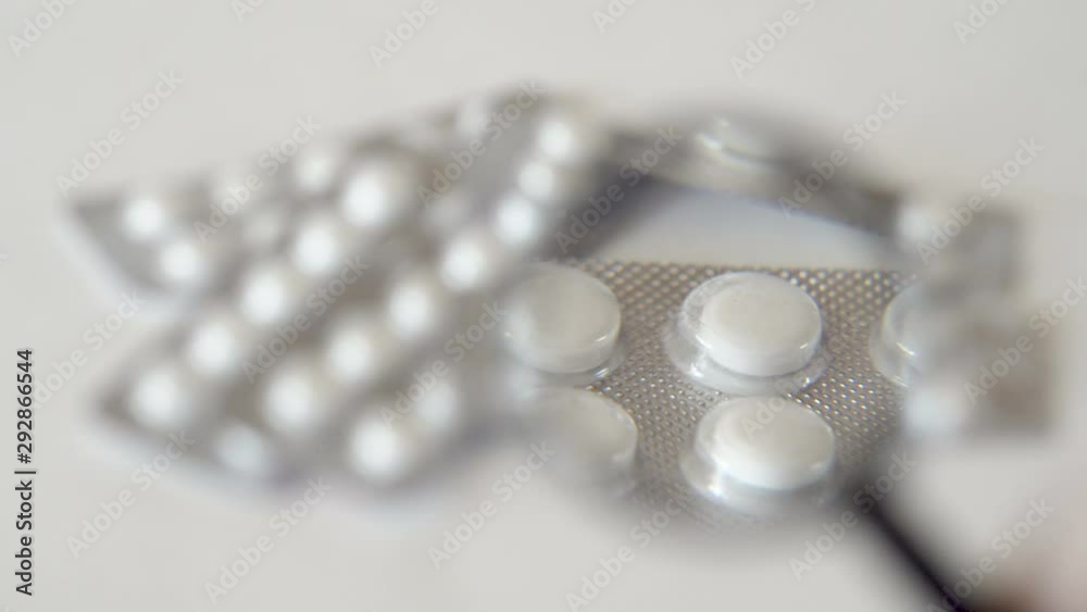 White round tablets in packs. Pills lie on a white surface. A hand in a white glove holds a magnifying glass. Looking through the glass. Medical product. The cure for the disease. Close-up.