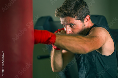 lifestyle gym portrait of young attractive and fierce looking man training boxing at fitness club doing heavy bag punching workout with wrist wraps in badass fighter look