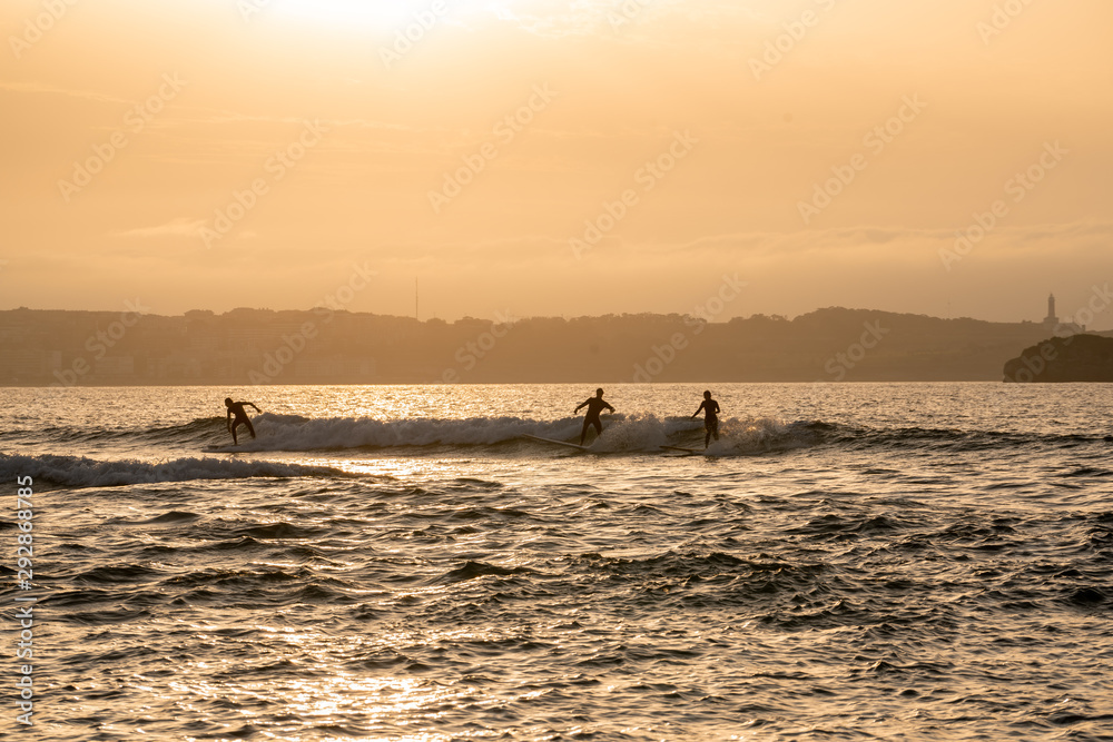 Fototapeta premium Wide image of surfers silhouettes surfing and riding waves at sunset o sunrise