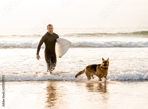 Canvas Print Surfer man with his dog german shepherd and surfboard playing and surfing on the