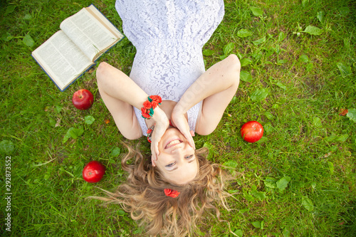 caucasian happy student girl in cute white dress rest on the green summer grass with book and red apples © goldeneden